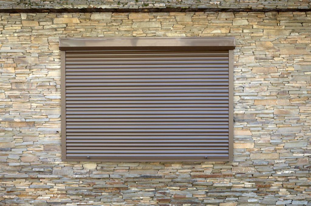 A window with brown metal rolling shutters. Wall decoration with artificial flatten stone